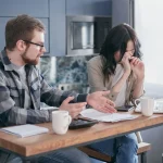 Persona preocupada mirando documentos financieros en una mesa de cocina modesta