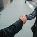Dos personas cerrando un acuerdo comercial con un apretón de manos junto a documentos en una mesa de oficina