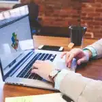 Hands adjusting budget sliders and cost metrics on a laptop screen in an office setting