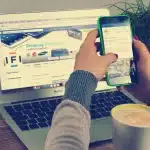 Hands typing on a laptop showing a company webpage with checklist and sticky notes in an office